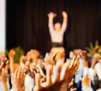 Alle Zuhörer strecken ihre Hände hoch um Moderator auf der Bühne nachzumachen. Dies soll als Symbolbild für eine agile Transformation dienen