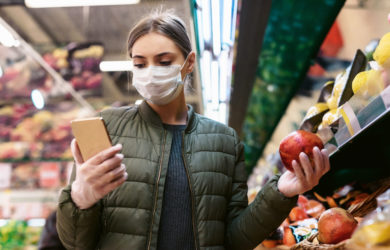 Eine junge Frau in einer Einwegmaske benutzt eine Inddor-Navigation und Tracking-App beim Einkaufen im Laden