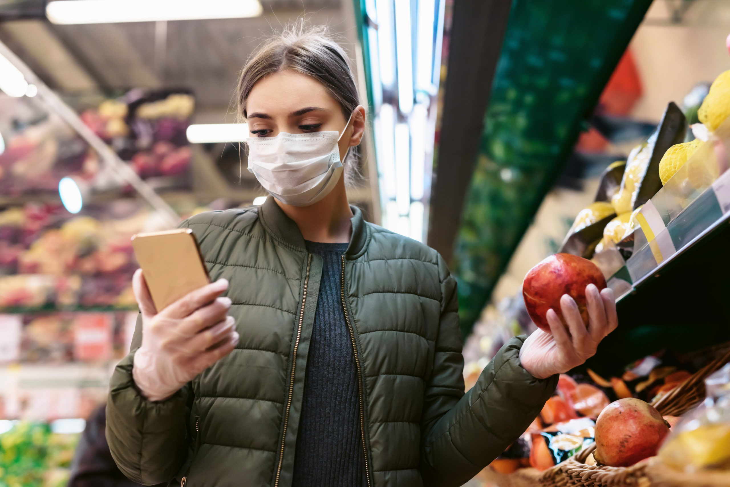 Eine junge Frau in einer Einwegmaske überprüft eine Einkaufsliste auf einem Smartphone im Supermarkt. Soziale Entfernung während der Pandemie des Coronavirus Covid-19