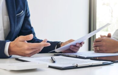 Zwei Geschäftspersonen im Gespräch, eine Person hält ein Dokument, auf dem Tisch liegen Unterlagen, ein Tablet und ein Kugelschreiber – symbolisch für Versicherungsberatung