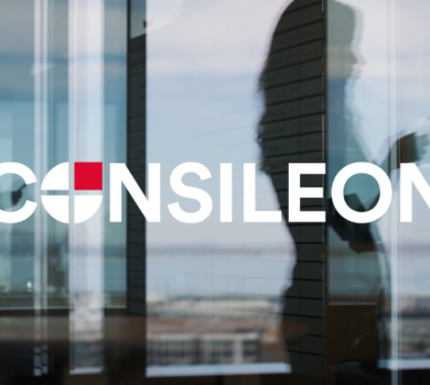 Frau steht am Fenster ihres Büros und schaut auf ihr Mobiltelefon, symbolisch für Kontaktaufnahme mit Consileon