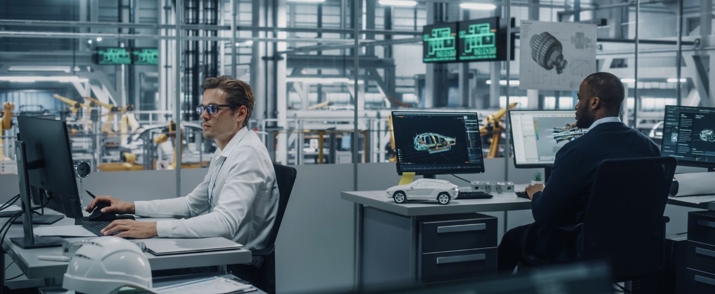 Zwei Ingenieure arbeiten an Computern in einer modernen Produktionshalle mit Robotern im Hintergrund und 3D-Modellen auf den Bildschirmen, symbolisch für digitale Entwicklung