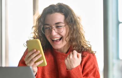 Eine junge Frau sitzt mit ihrem Smartphone in der Hand vor ihrem Laptop am Schreibtisch und wirkt fröhlich/aufgeregt.