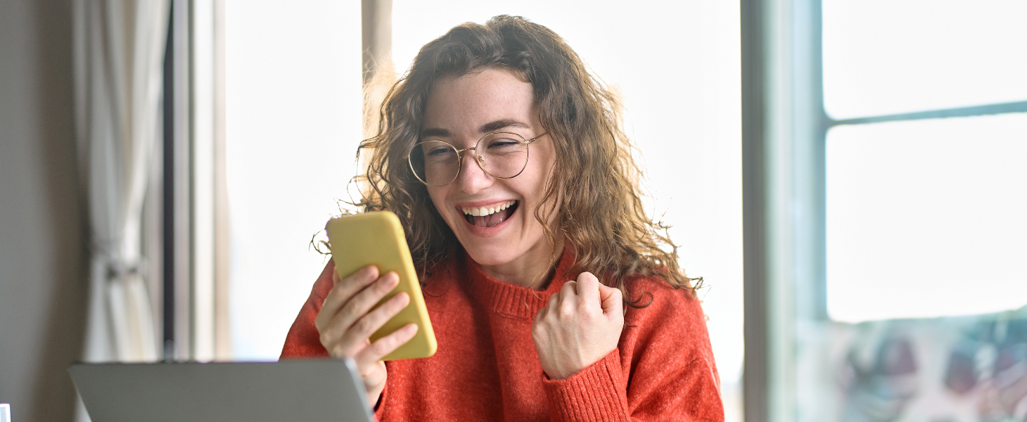 Eine junge Frau sitzt mit ihrem Smartphone in der Hand vor ihrem Laptop am Schreibtisch und wirkt fröhlich/aufgeregt.