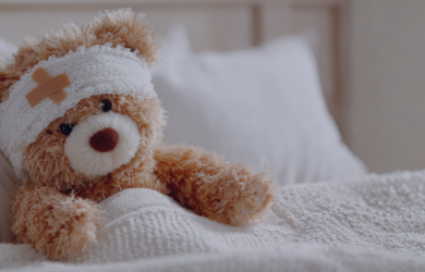 Ein Teddybär mit Kopfverband sitzt zugedeckt in einem Bett.