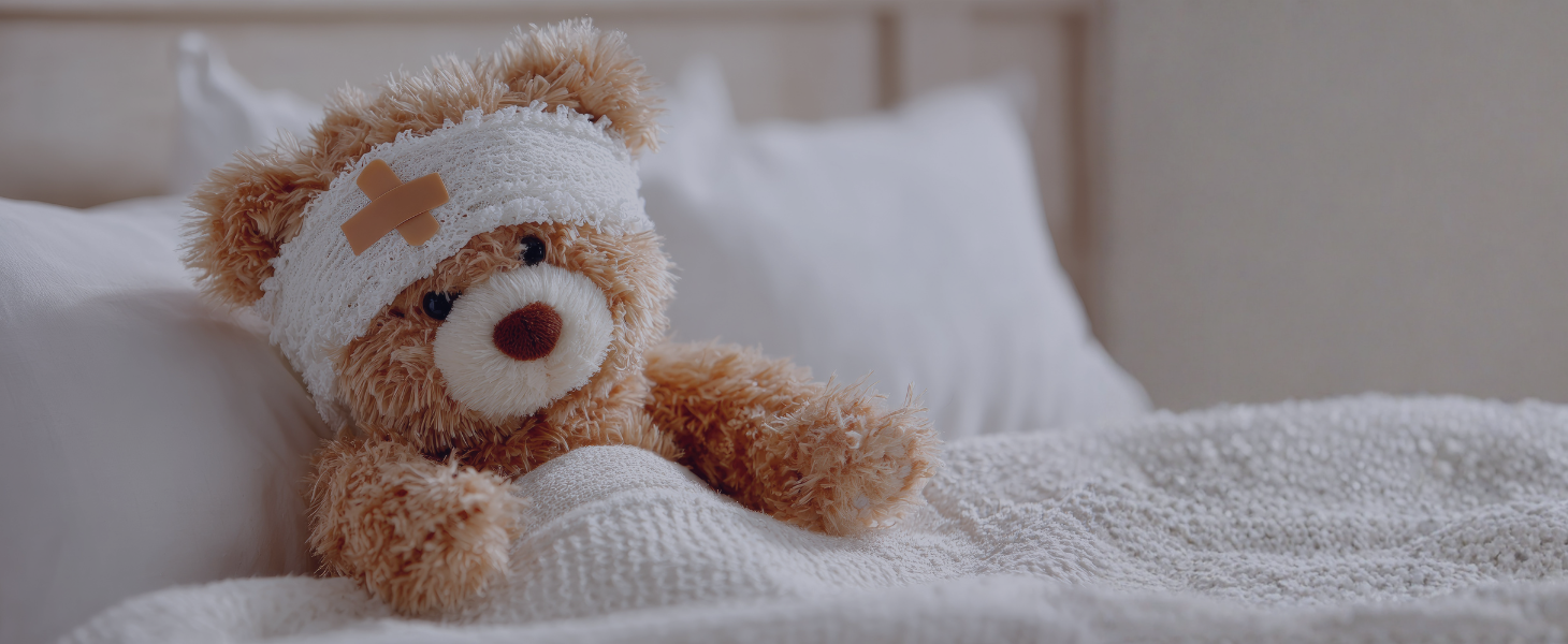 Ein Teddybär mit Kopfverband sitzt zugedeckt in einem Bett.